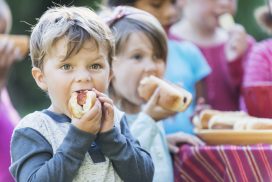 A little boy, 4 years old, eating a hotdog at a picnic.  His mouth is full as he takes a bite.  He is wearing a gray shirt with blue sleeves. Standing behind him are other children eating hotdogs.