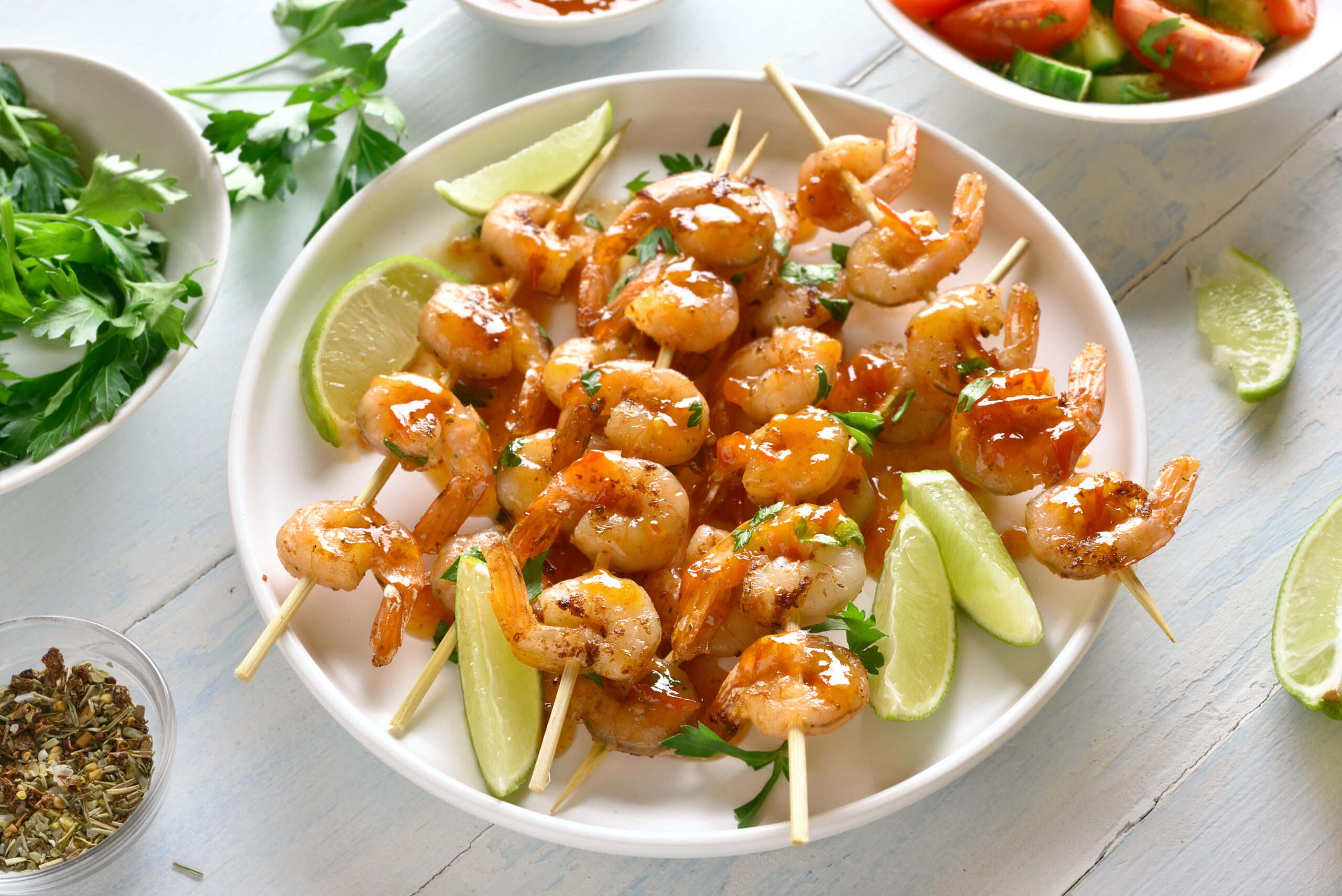 Prawns skewers with greens, spices, lime and sauce on white plate over wooden table. Grilled shrimp skewers. Tasty seafood.