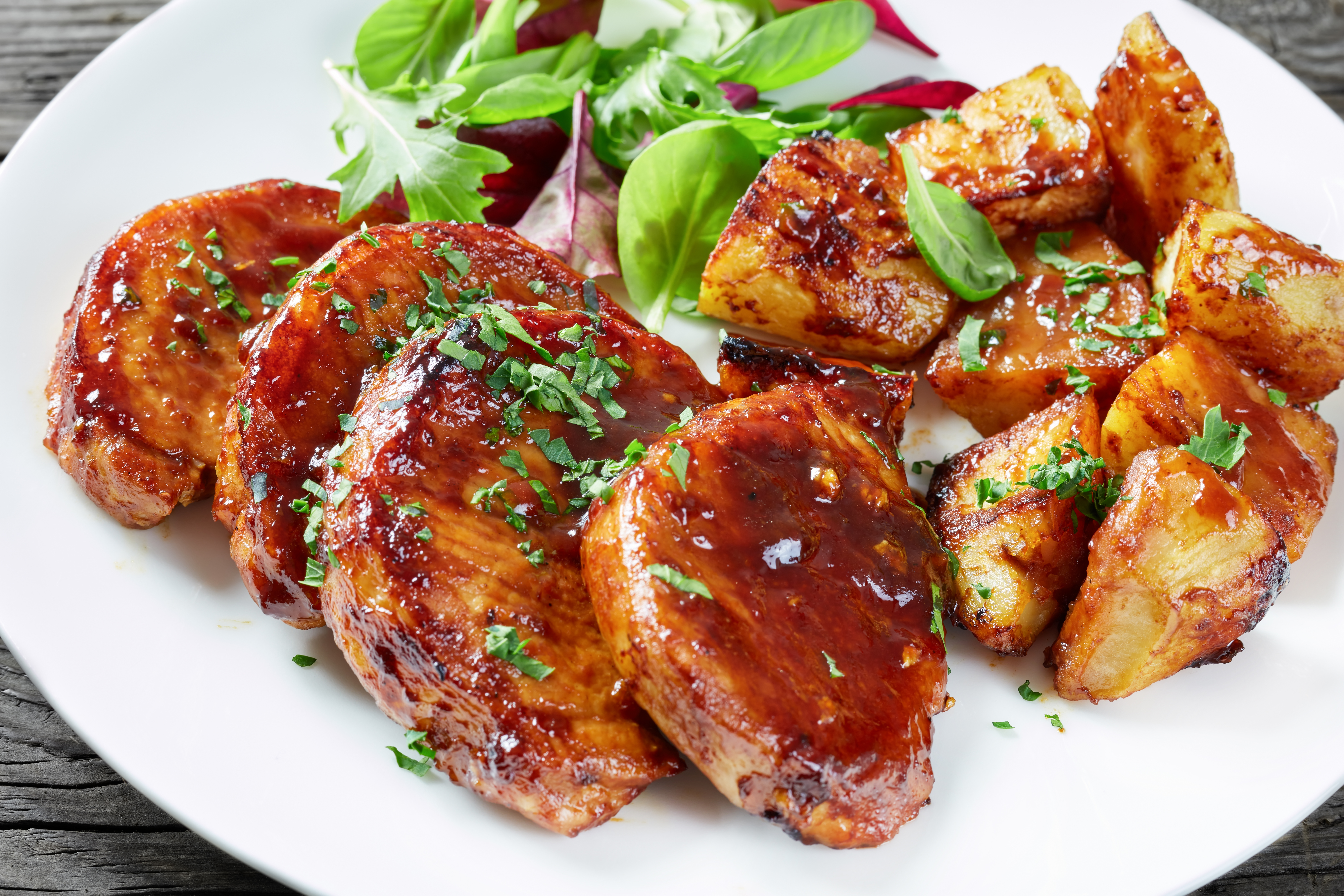 close-up of Oven Baked sticky glazed Pork Chops with baked Potatoes served with fresh green salad on a white plate on a rustic table, view from above