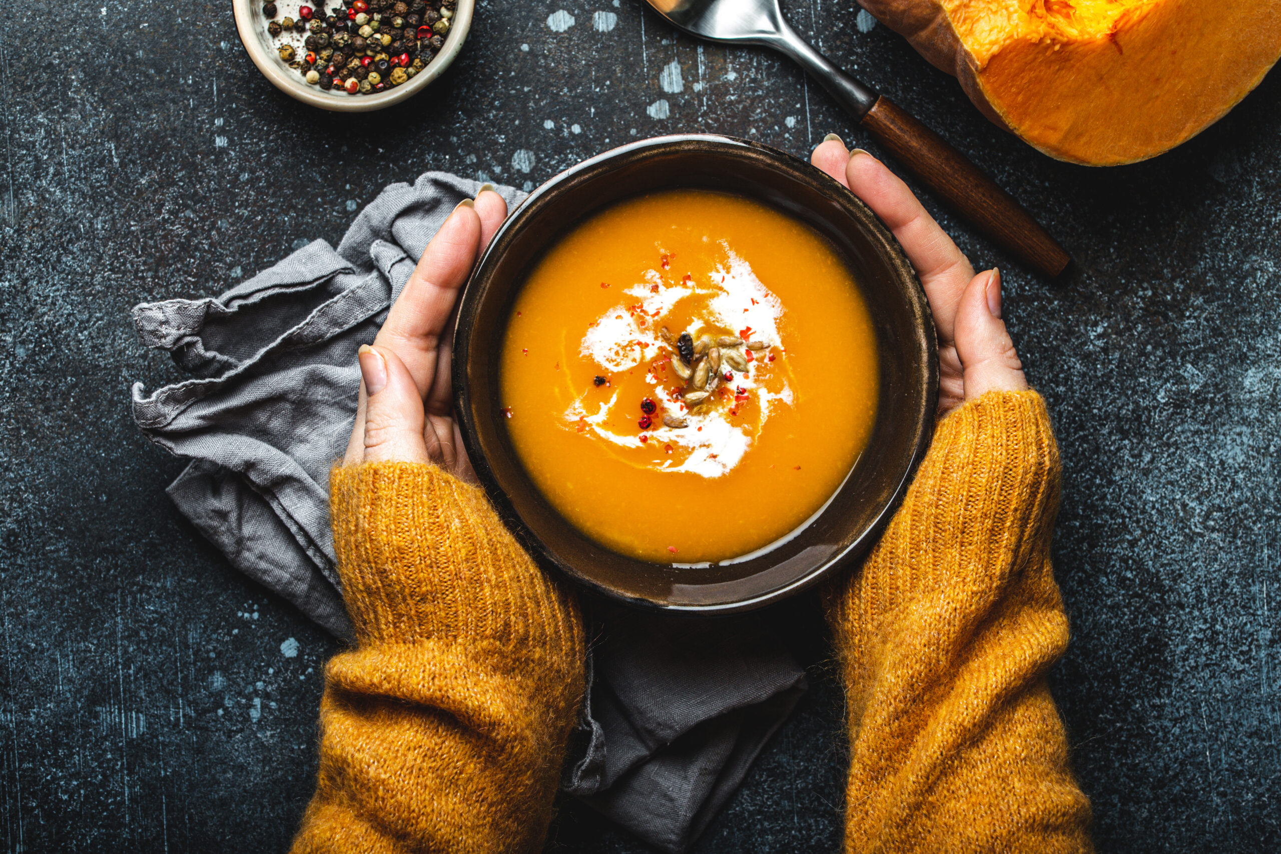 Female hands in yellow knitted sweater holding a bowl with pumpkin cream soup on dark stone background with spoon decorated with cut fresh pumpkin, top view. Autumn cozy dinner concept